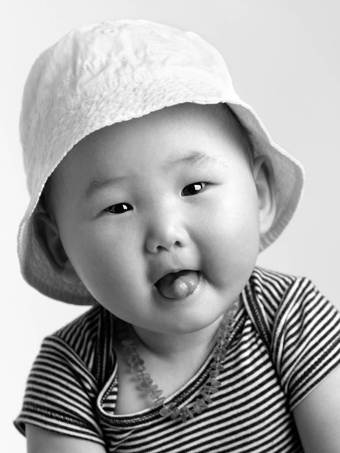 A black and white photo of a smiling baby wearing a striped shirt and a sun hat, playfully sticking out their tongue. The mood is joyful and playful.