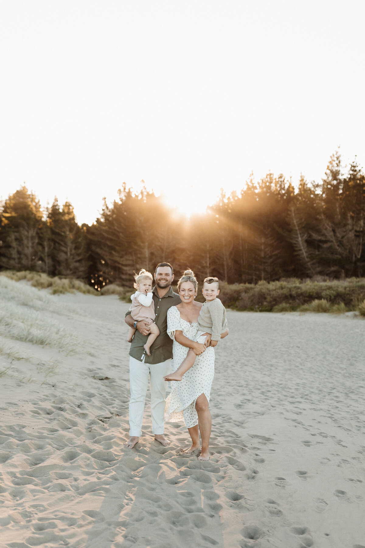 A mother and father and two little children smile at the camera at the beach while the sunsets in the background during their family photoshoot in christchurch