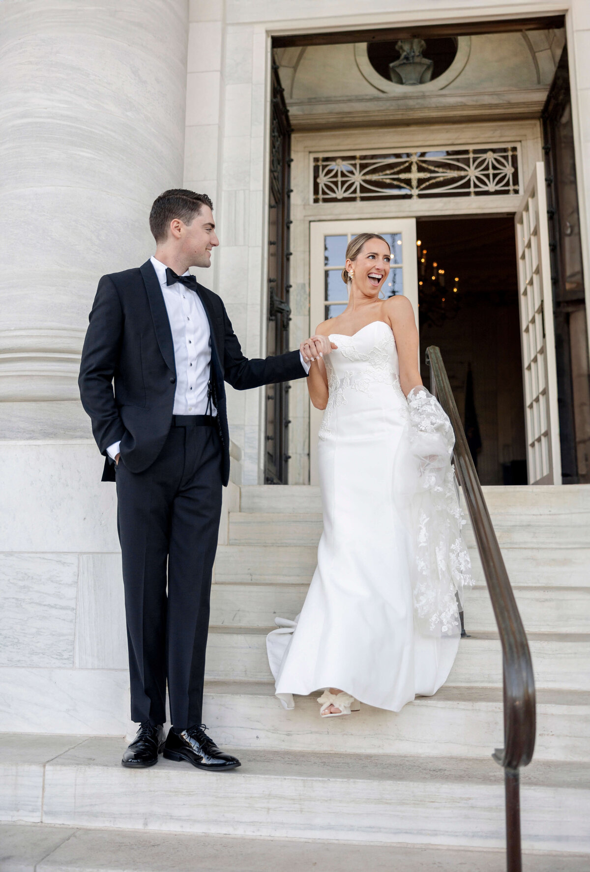 A couple descends stone steps outside a building. The groom wears a black tuxedo, while the bride is in a white wedding gown and holds lace fabric. They appear joyful and are clasping hands.