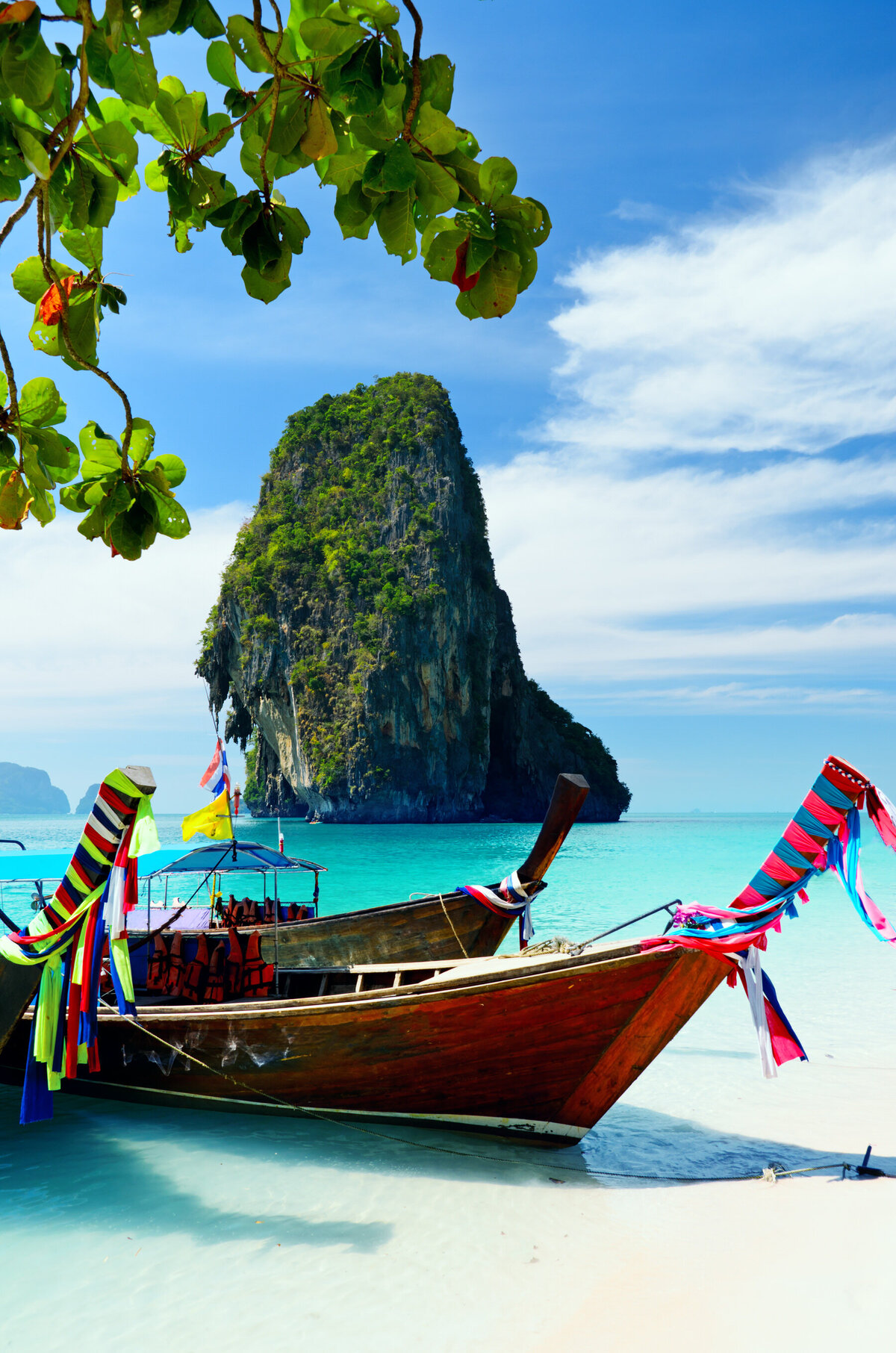 A wooden long-tail boat adorned with colorful ribbons is moored on a sandy beach with clear turquoise water. A lush, tree-covered limestone rock formation rises in the background under a blue sky with scattered clouds. Green leaves frame the scene.