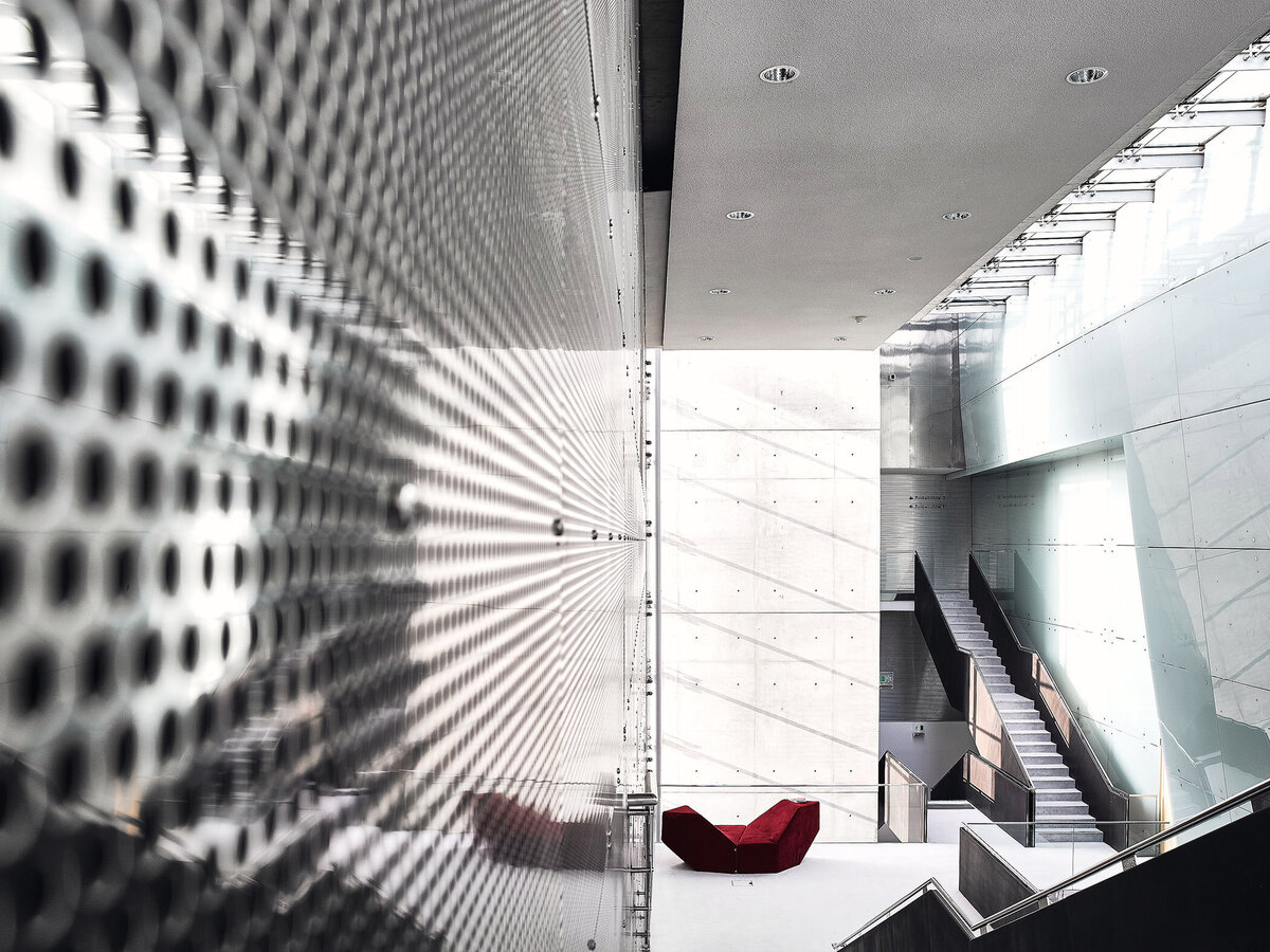 Architekturfotografie eines lichtdurchfluteten Innenraums mit Glasfassade, Betonwänden und markantem roten Sofa – moderner Baukörper mit klaren Linien und starker Lichtführung, fotografiert von Atelier Schulte in Wien.