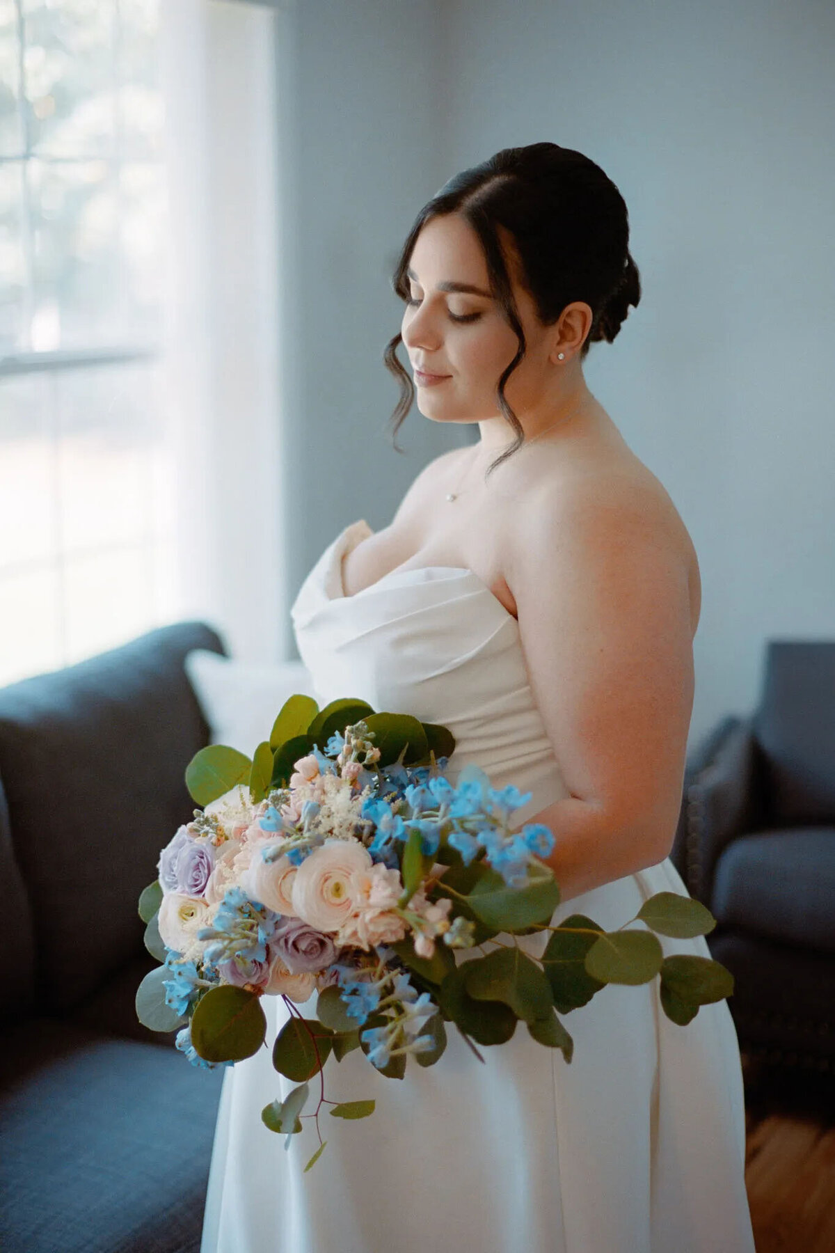 A bride in a white strapless gown holds a pastel bouquet with blue and pink flowers, standing indoors by a window with soft natural light, eyes closed and hair in an elegant updo—captured beautifully by an NJ wedding photographer.
