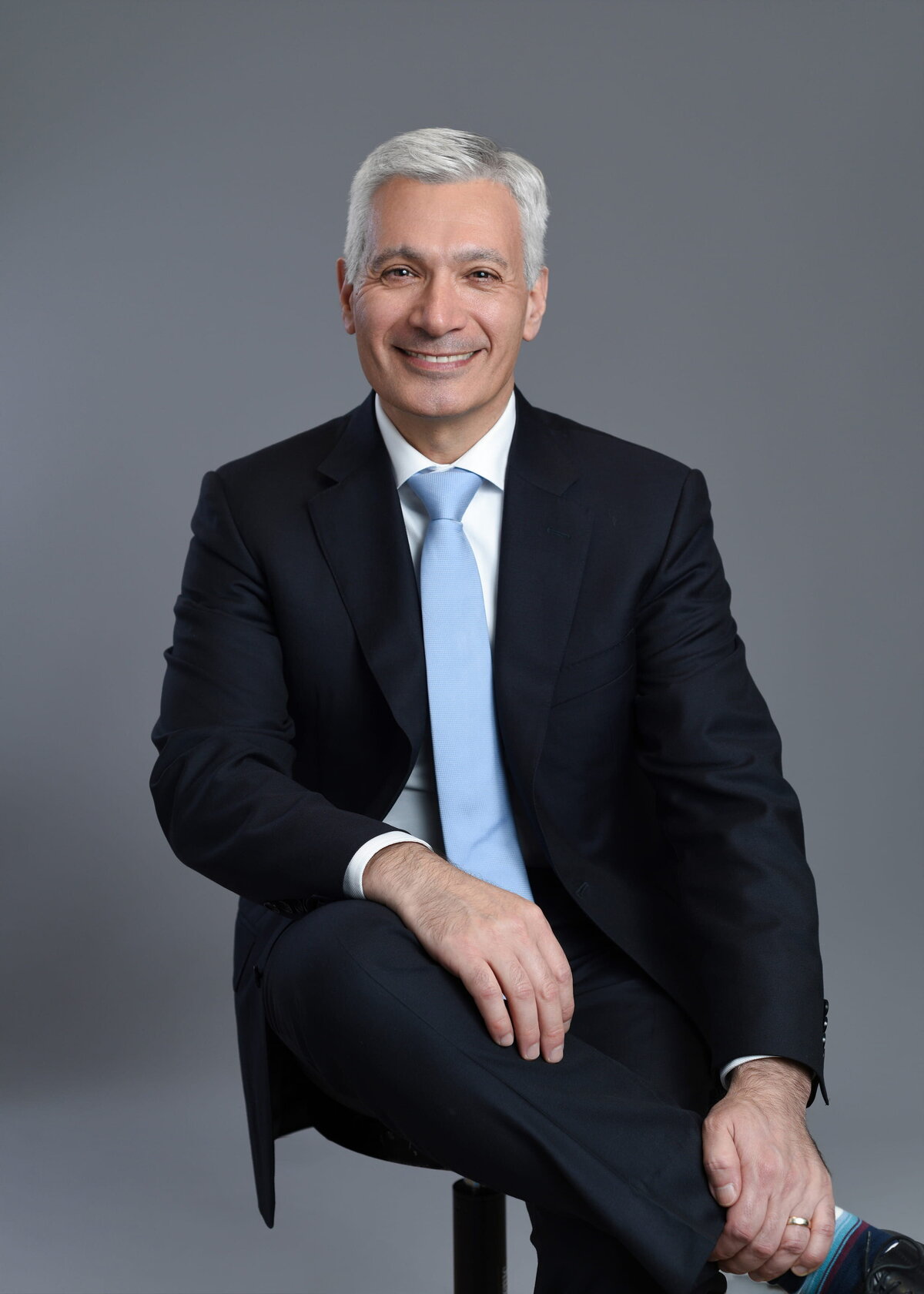 Professional headshot of a distinguished individual seated against a neutral gray backdrop, exuding confidence and approachability. Ideal for business and personal branding photography