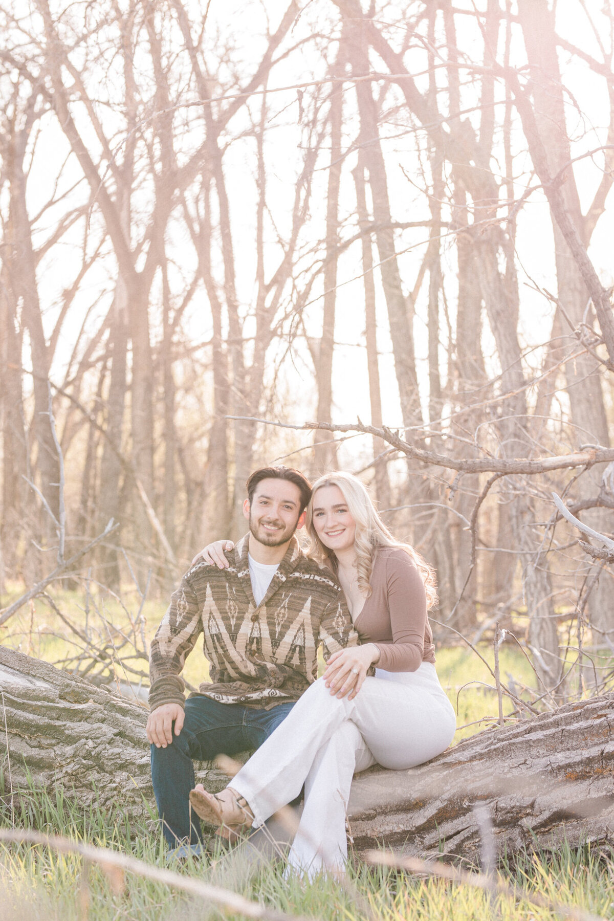 A blond woman and dark haired man sit on a fallen tree and smile brightly for the camera at Walden Pond in Boulder, Colorado