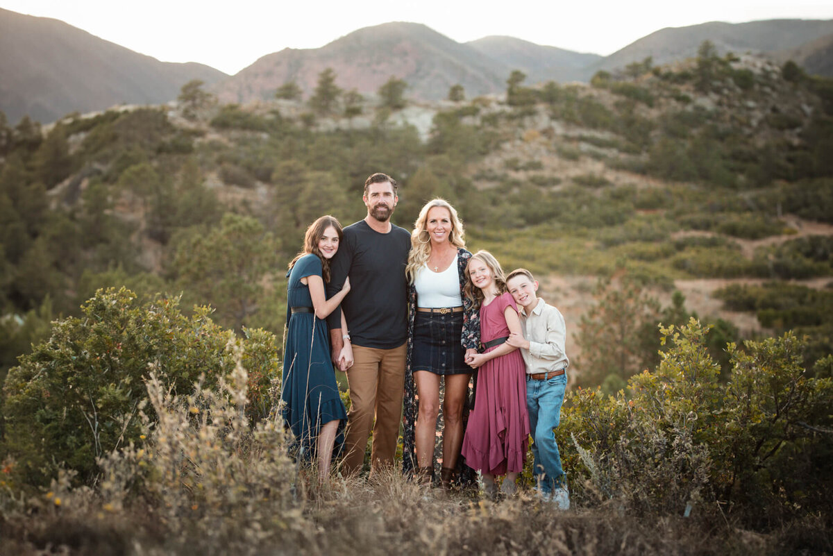 Three children hug onto mom and dad as they smile together in a mountain trail
