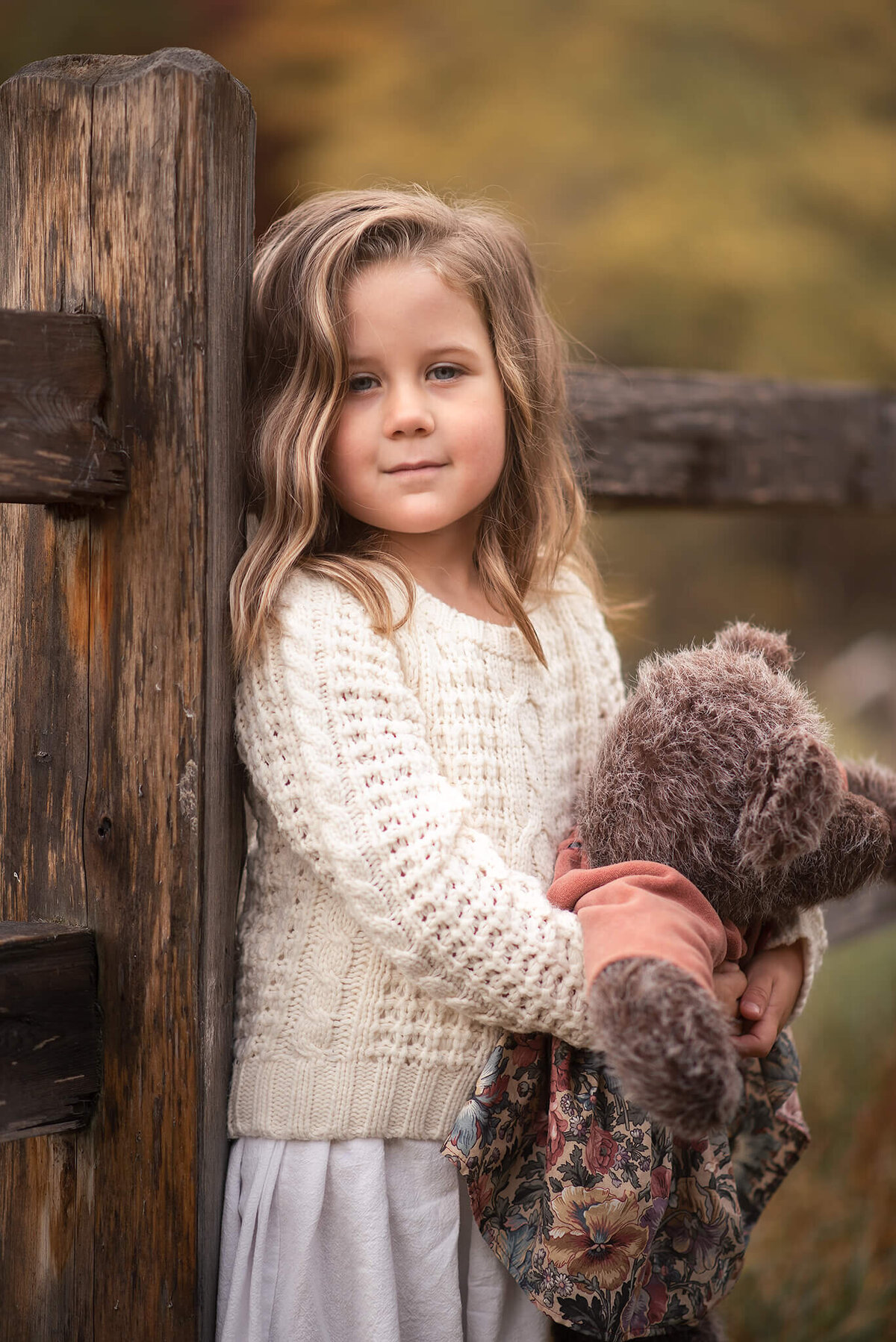A beautiful little girl with blonde hair wears a white knit sweater while holding a teddy bear. She is leaned up against a fence in a park in Kamloops.
