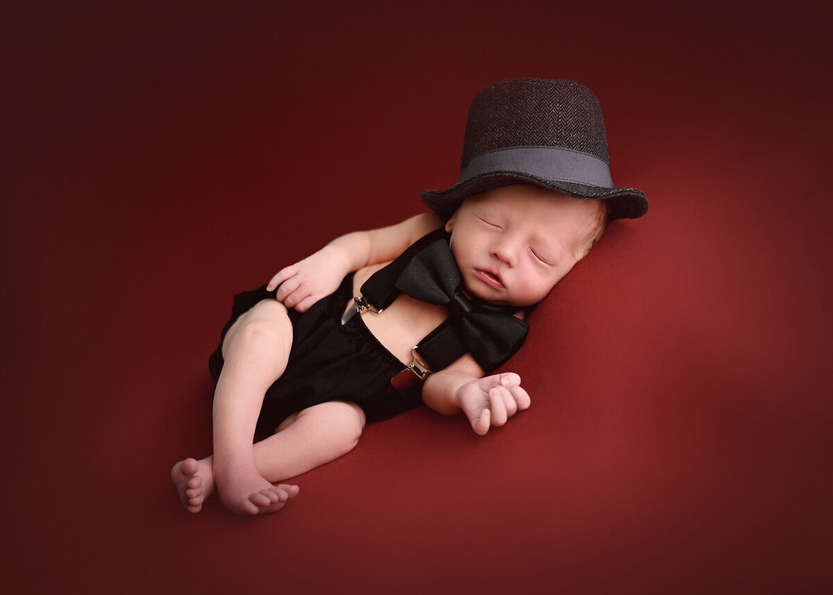 A newborn baby wearing a hat and bow tie sleeps on a red background