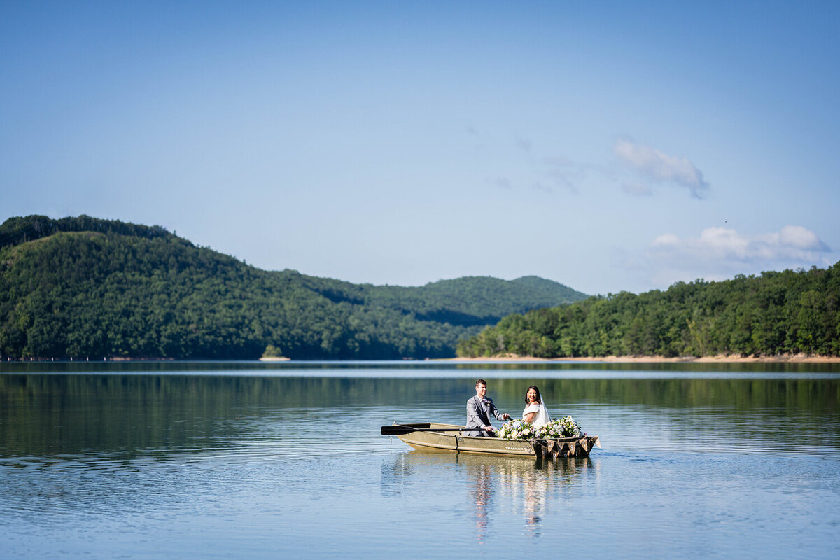 A bride and groom sit in a rowboat in the waters of Carvin’s Cove with the rolling hills of Southwest Virginia in the background. The boat is adorned with pastel flowers and a sign that reads “just married” on the back of the boat. The groom is moving the oars while the bride looks back towards the shore to smile.