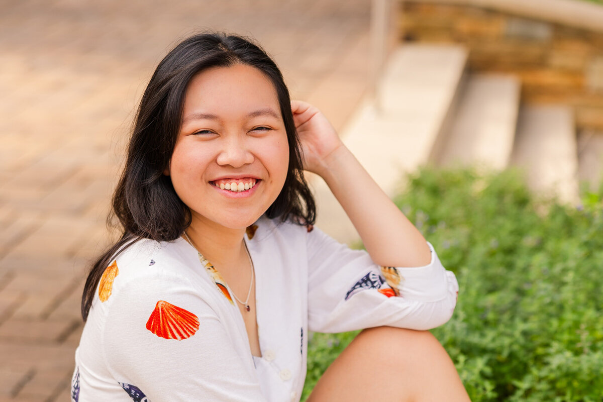 teenager girl sitting and laughing at the camera with one hand in her hairs with a city background in Sandy spring during photoshoot