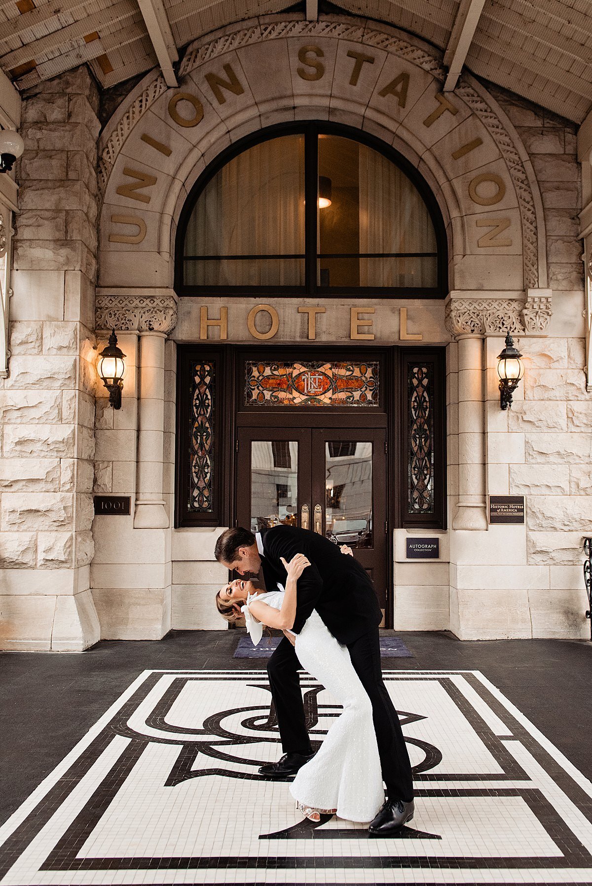 The groom dip the bride as he kisses her in front of the main entrance of the Union Station Hotel in Nashville. The bride is wearing a floor length  fitted sheath dress with a ruffle over the shoulder. The groom is wearing a black tuxedo  with a white shirt. They are standing on the mosaic floor in front of the hotel. Featured is a large white rectangle bordered in black with USH written out as a monogram for the hotel.  Behind them is the rough hewn stone historic train station in downtown Nashville. The  door frame is dark wood with stained glass windows and transoms. On either side of the doors are two round sconces lit up. Above the door is a large half oval window with a curtain drawn in front of it. The words Union Station Hotel is detailed around the arch of the window in large gold lettering.