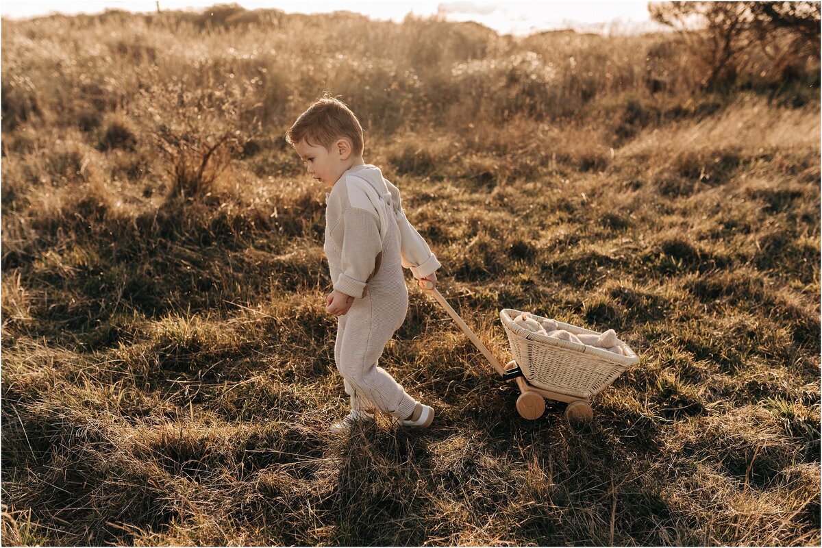 boy in jamie kay clothing pulling maileg wicker trolley in long grass at sunset
