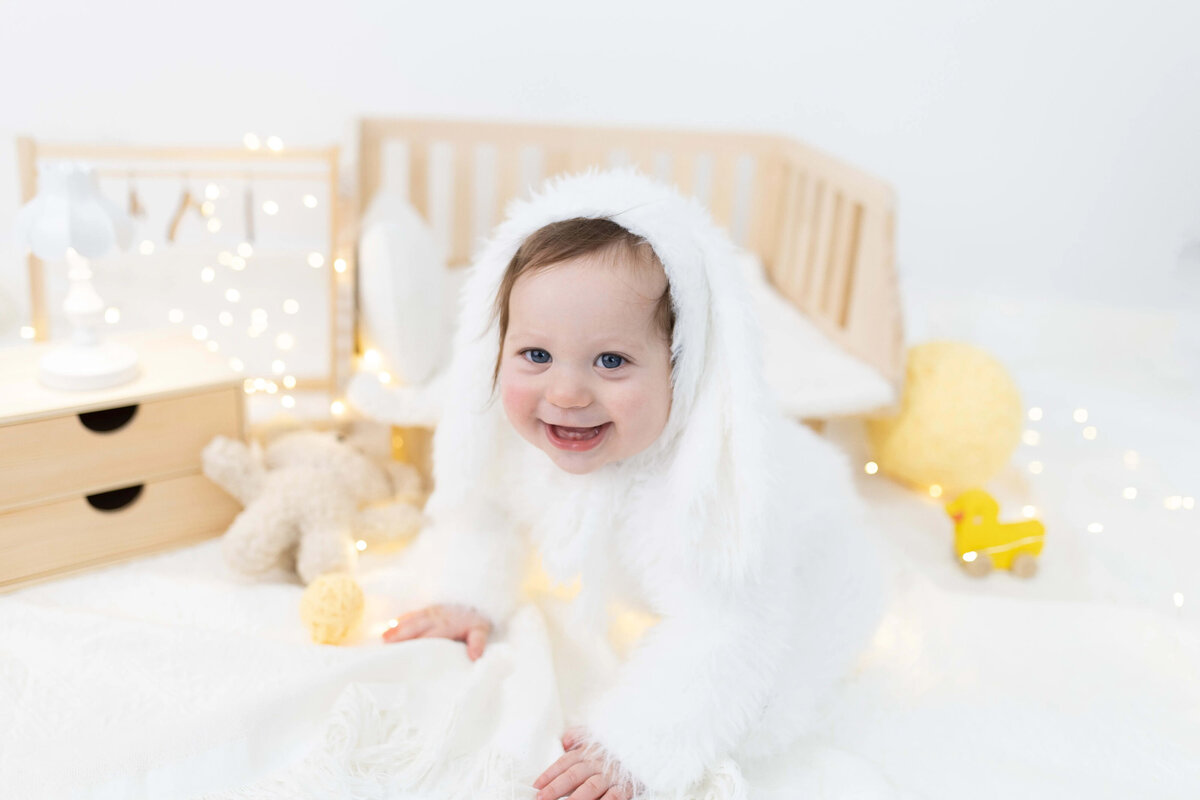 Smiling baby in white fuzzy outfit sitting on a mini wooden bed - styled milestone photography session in Maple Grove, MN