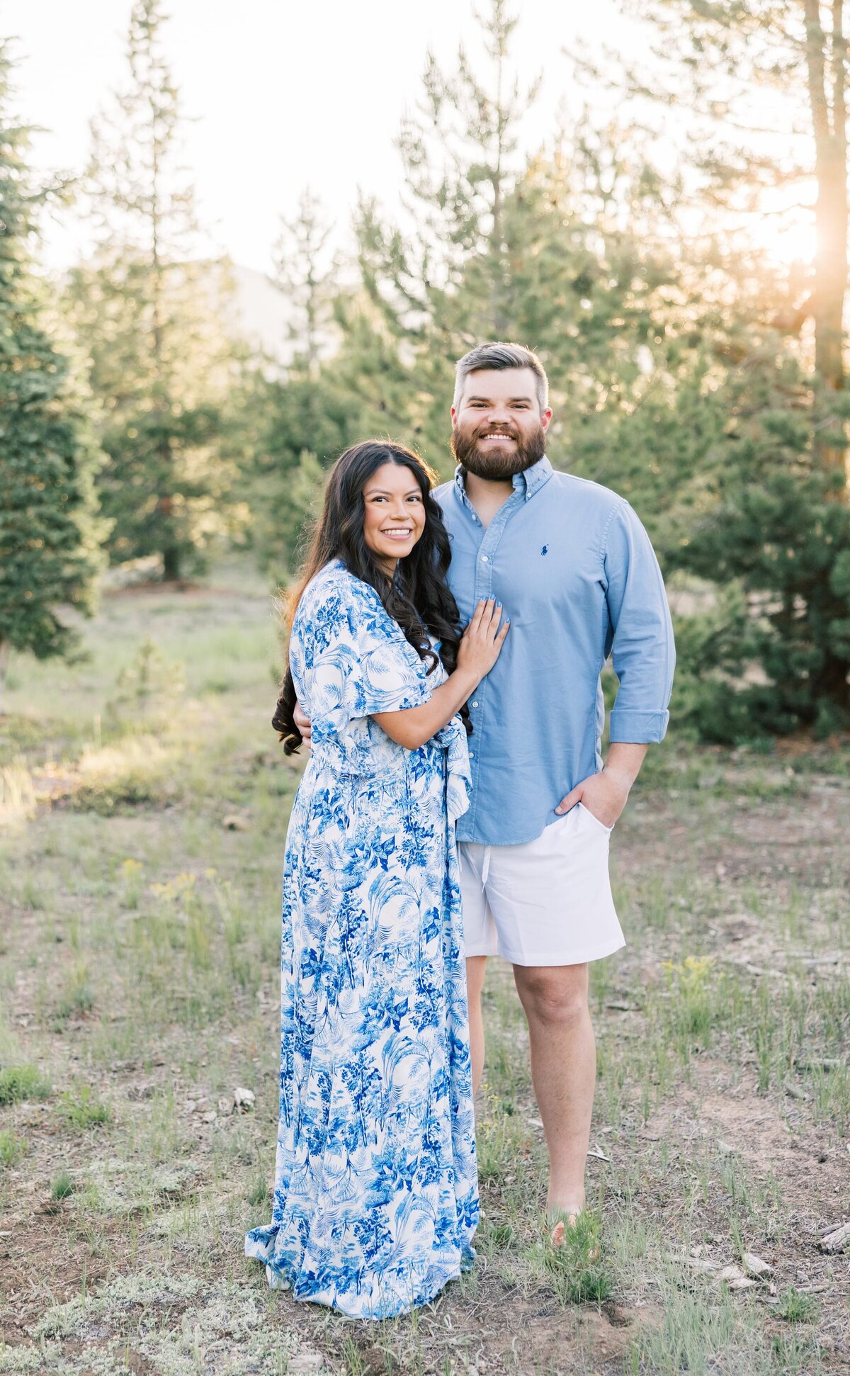This is a nice posed portrait of a young girlfriend and boyfriend. They are standing amongst the pine trees and her arm is on his chest. They are smiling at the camera. 