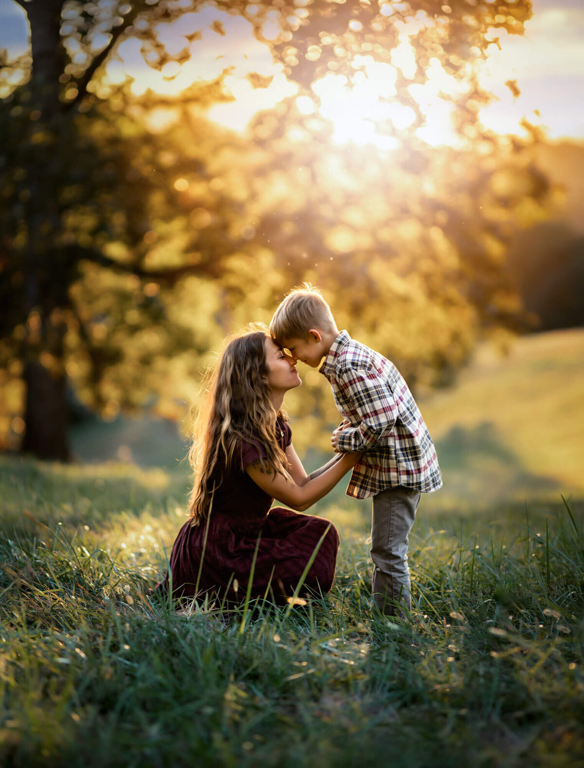 A mom in a maroon dress touching foreheads with her son while kneeling in the grass at sunset