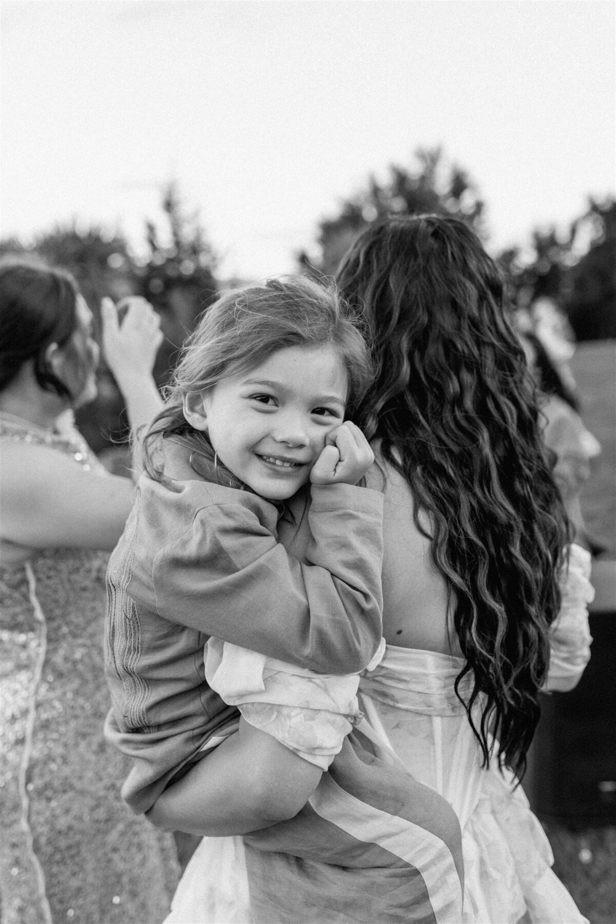 Black and white candid of a young boy smiling sweetly while being carried, nestled into the shoulder of a woman in a wedding gown with long wavy hair.