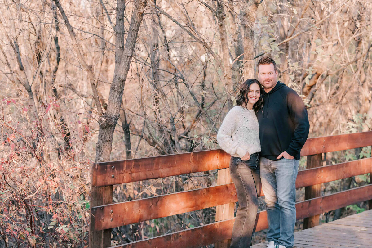 Mom and dad stand together by a wood railing on a small walking bridge in Saskatoon. 