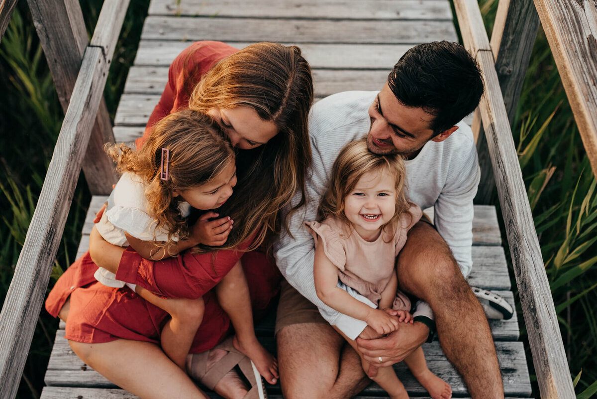 A young family consisting of a mother and father and two young girls snuggle together on a boardwalk during their photoshoot with Allison Wolf, Boston Family Photographer