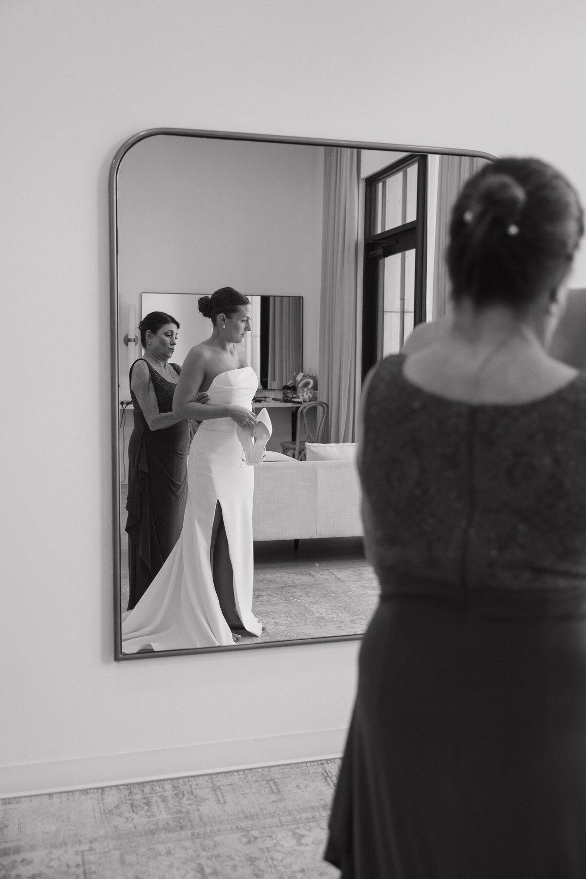 A mother helps zip up a wedding gown on her daughter in the reflection of a large mirror hanging on the wall. The dresses is strapless, white and satin. The bride holds pointed, closed toed heels in one hand and looks out the window. 