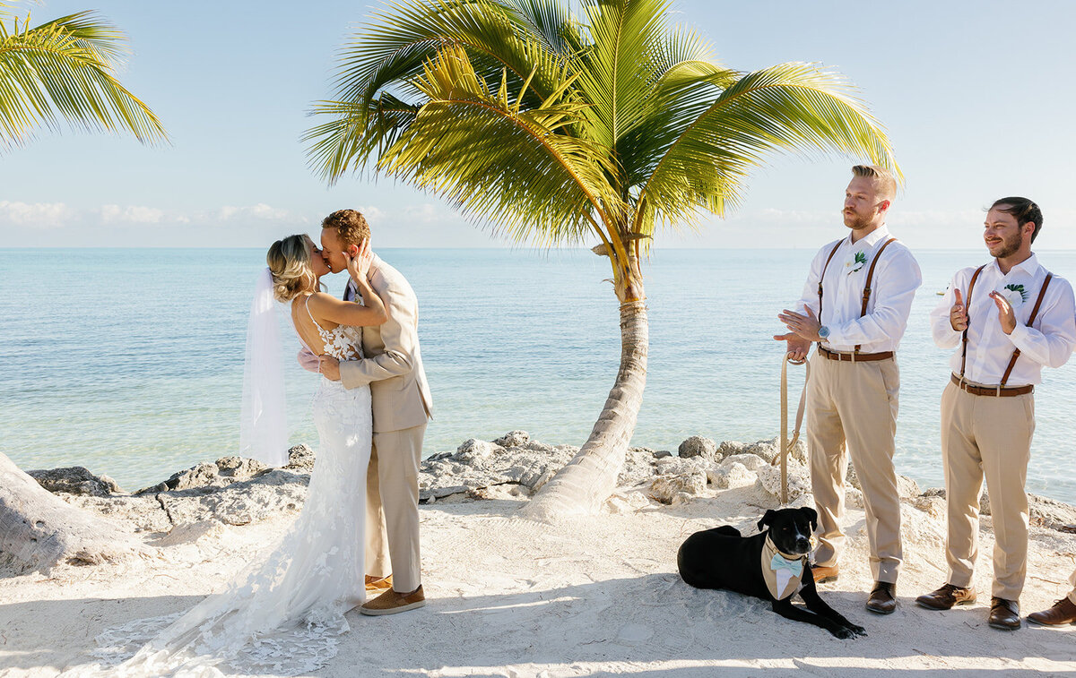 Ashley and Hunter share their first kiss as newlyweds with palm trees and the ocean behind them.