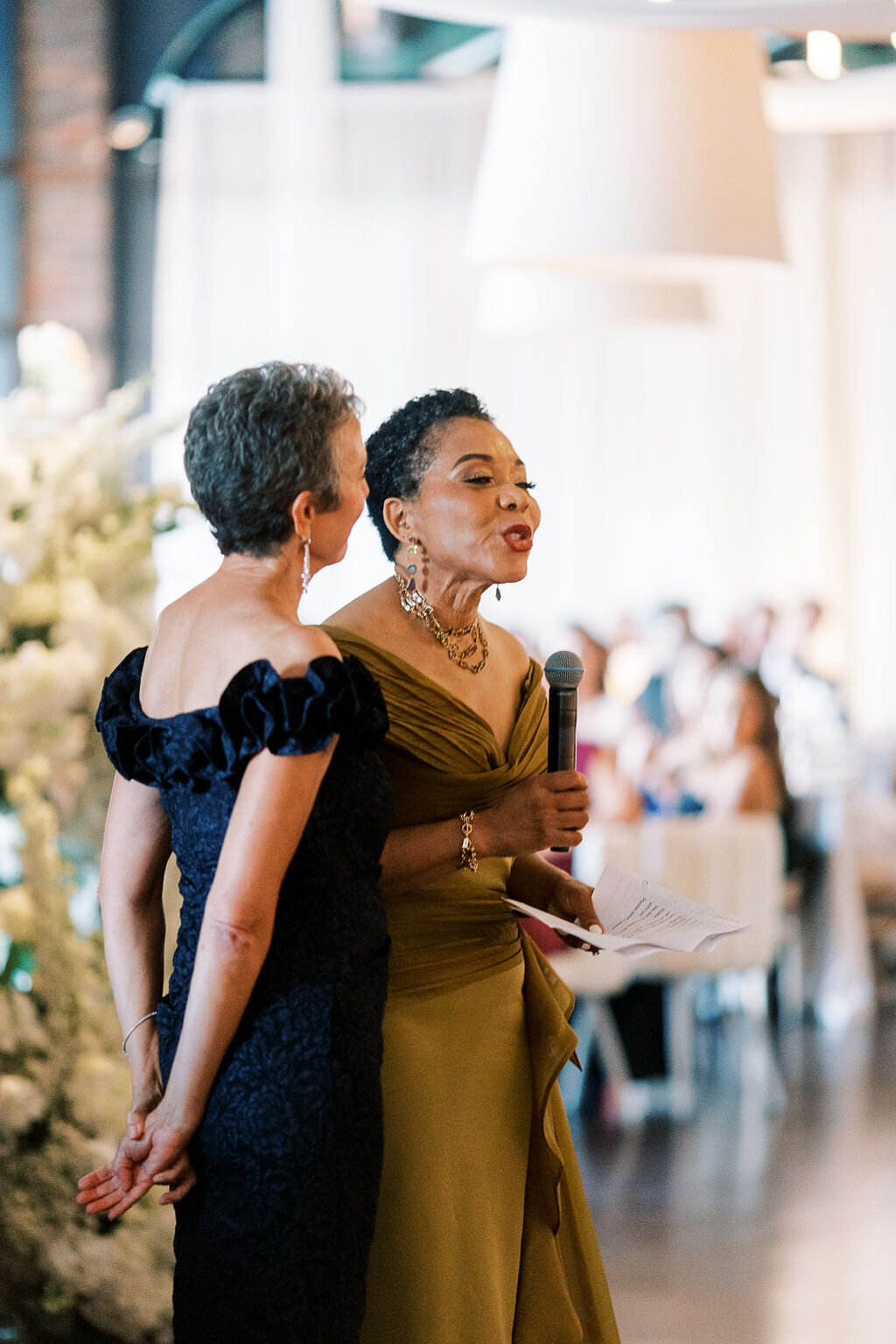 Two elegantly dressed women give a heartfelt speech at the reception, standing near beautifully arranged florals.