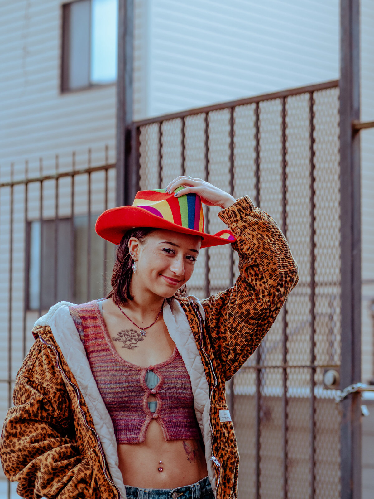 Creative poses in a rainbow cowboy hat and leopard jacket in front of a city fence – playful, queer-friendly brand portrait by Mewphotovideo in Austin, Texas