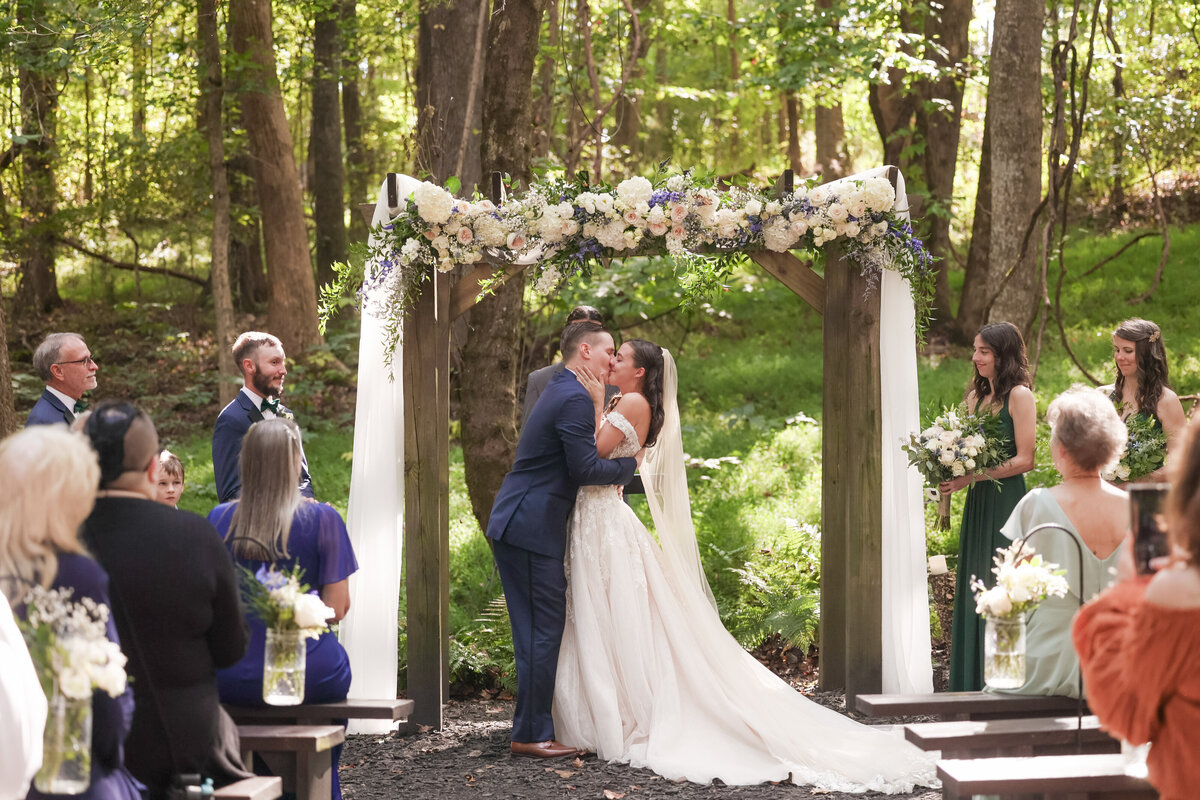wedding couple kissing under wooden arbor with colorful flowers with family and friends looking by Amanda Richardson Photography at Correy House in Union Point Georgia