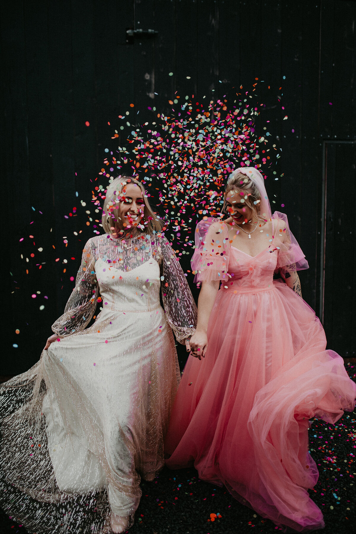 Our same sex couple throw colourful confetti outside the giraffe shed. the confetti and the girls pinks and white dresses contrast against the industrial black barn of the wedding venue.