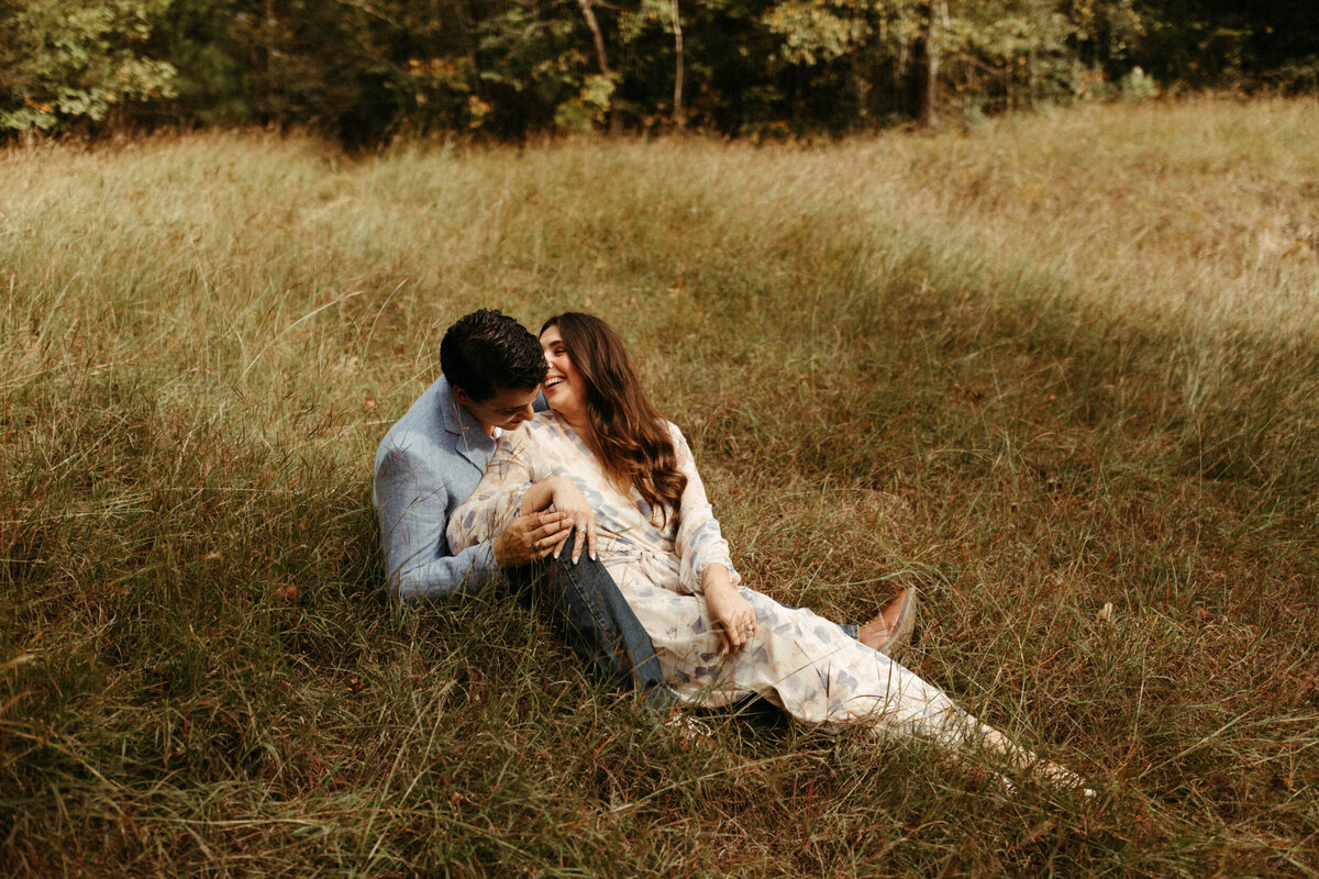A girl is sitting on the ground between her fiancé's legs in a grassy field and laughing.