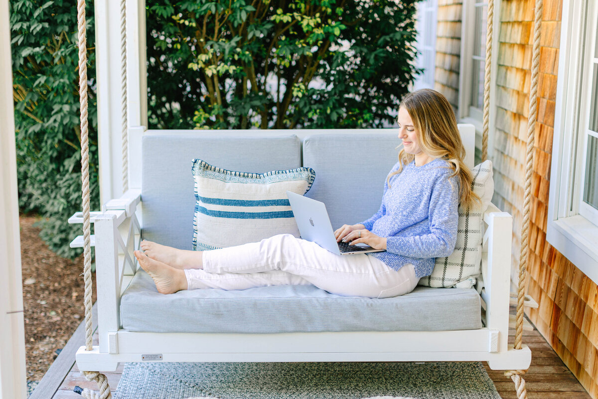 A woman in a cozy blue sweater and white pants lounges barefoot on a cushioned porch swing, working on her laptop. The swing is decorated with striped and plaid pillows, set against a backdrop of lush greenery and a shingled house exterior.