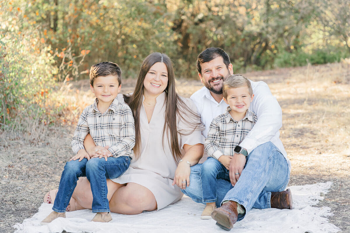 a family of 4 with two young boys sits on a white blanket at an Austin park and poses for their family photos.