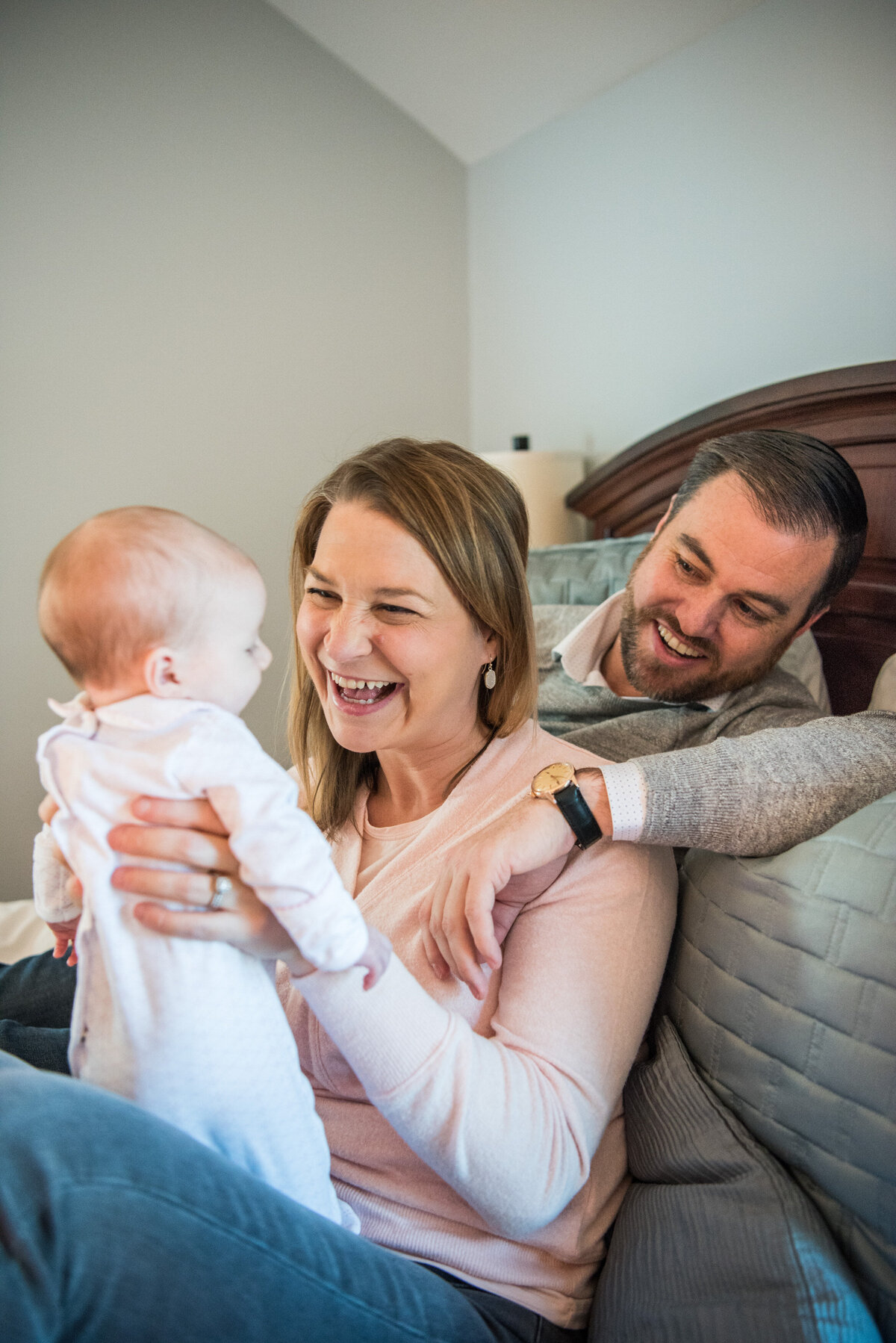 Two parents laugh with their newborn while sitting on a bed.