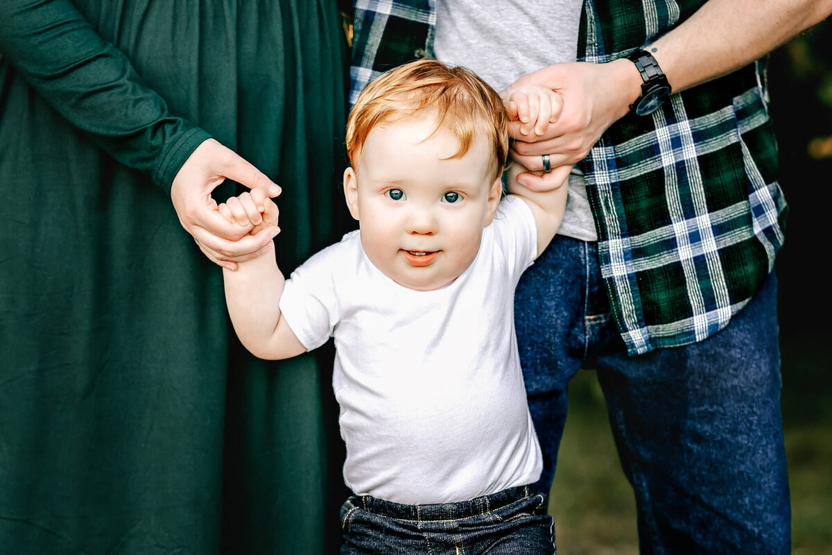 A toddler with red hair and a white shirt holds hands with two adults. One sports a green dress, the other a gray shirt with a green flannel. Captured outdoors on grass by Bloomington family photographer, this image truly reflects incredible moments in studio and outdoor portraits.