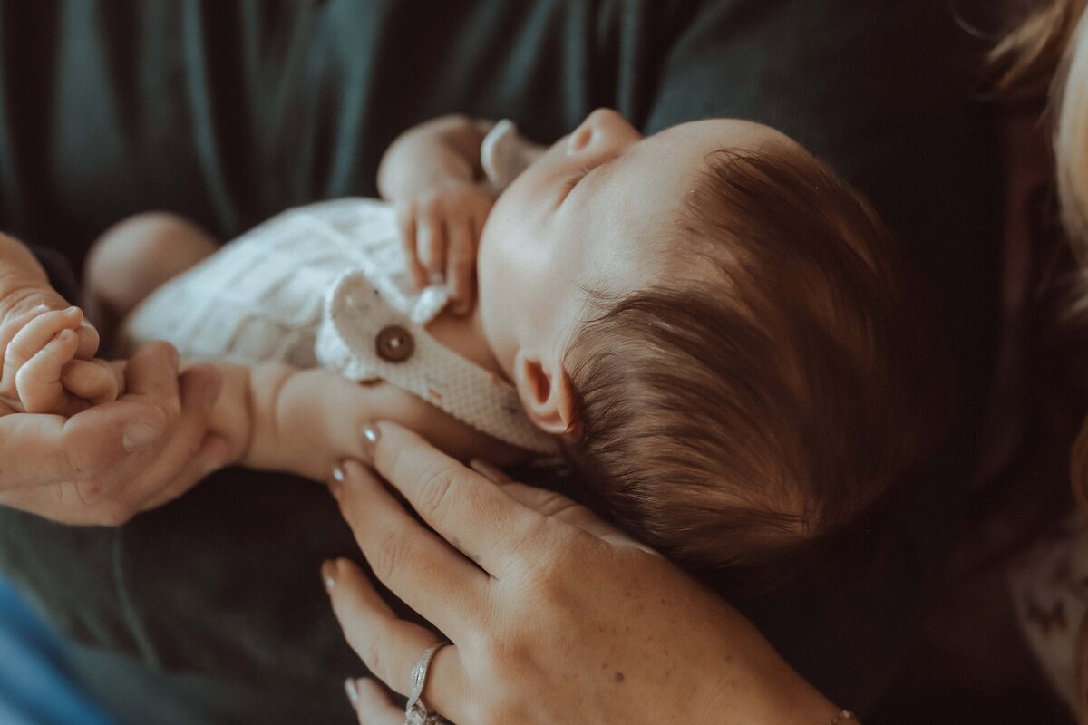 A baby wearing a white outfit lies asleep, cradled in an adult's arms. Hands gently hold the baby's arm and head.