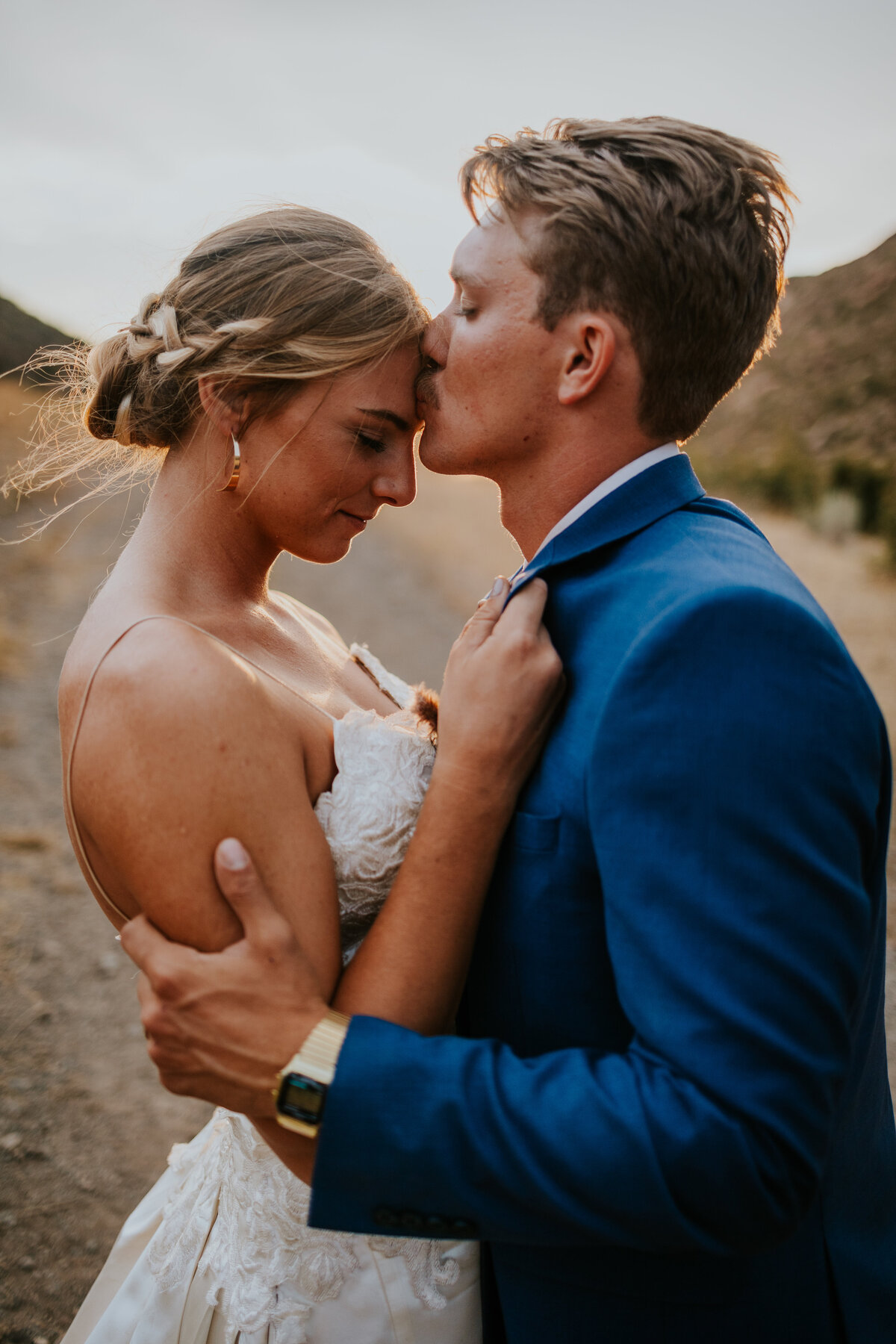 Groom kisses brides forehead while she holds his collar as they stand in front of sunset.