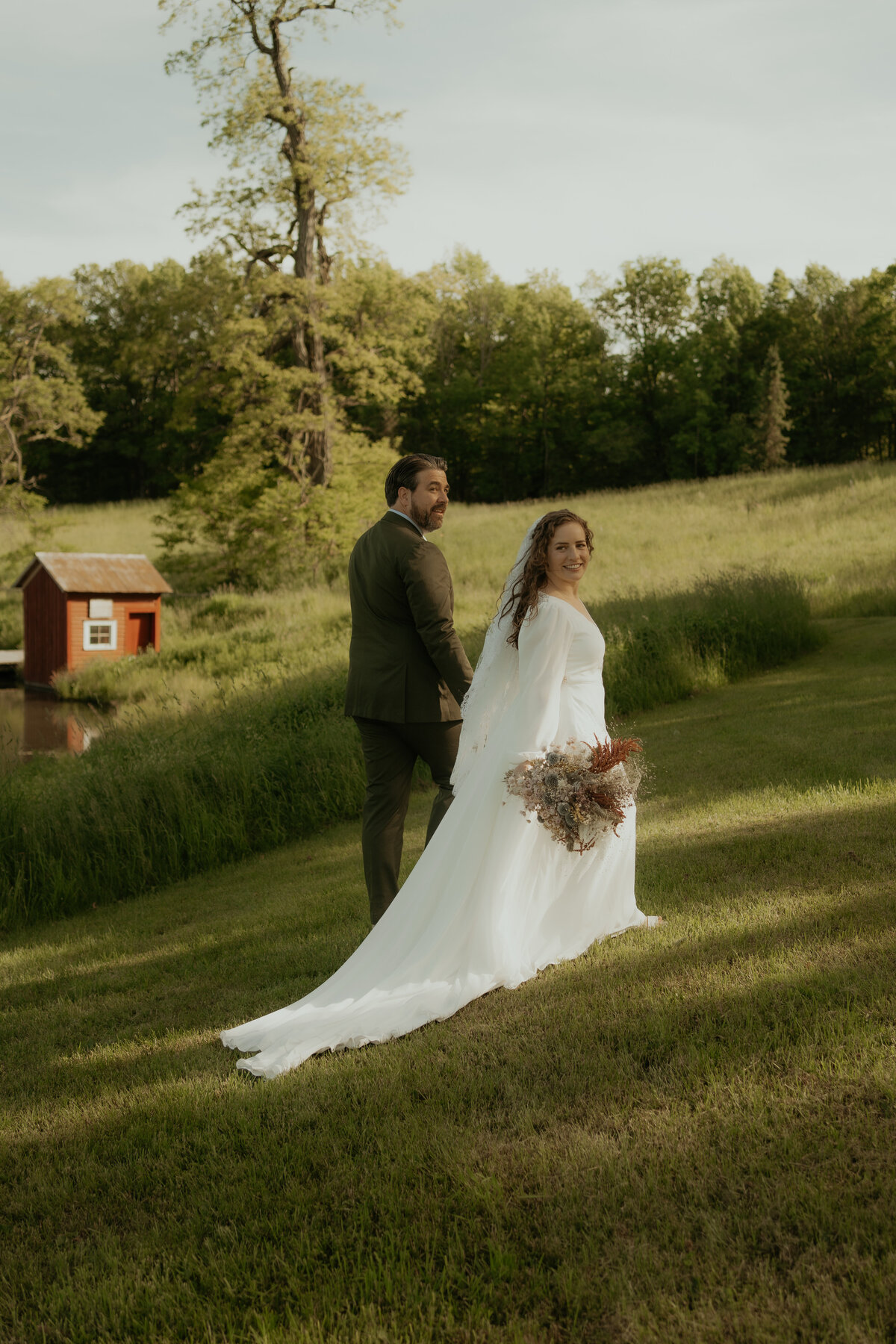 A bride and groom walk hand in hand on a lush green hillside at their wedding at Blenheim Hill Farm in Jefferson, NY with a small red barn in the background. The bride, in a flowing white gown, smiles back at the camera while holding a bouquet of dried flowers. The groom, in a dark suit, also looks back, sharing the moment. The setting is serene and picturesque, perfect for a documentary-style wedding photograph that captures the couple's natural joy and the beauty of their surroundings.