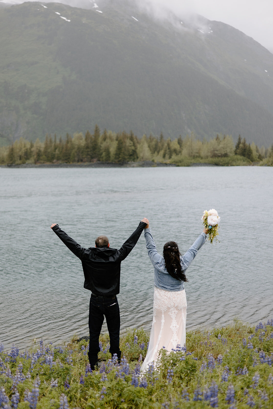 Elopement couple raises hands by misty lake and mountain in Alaska.