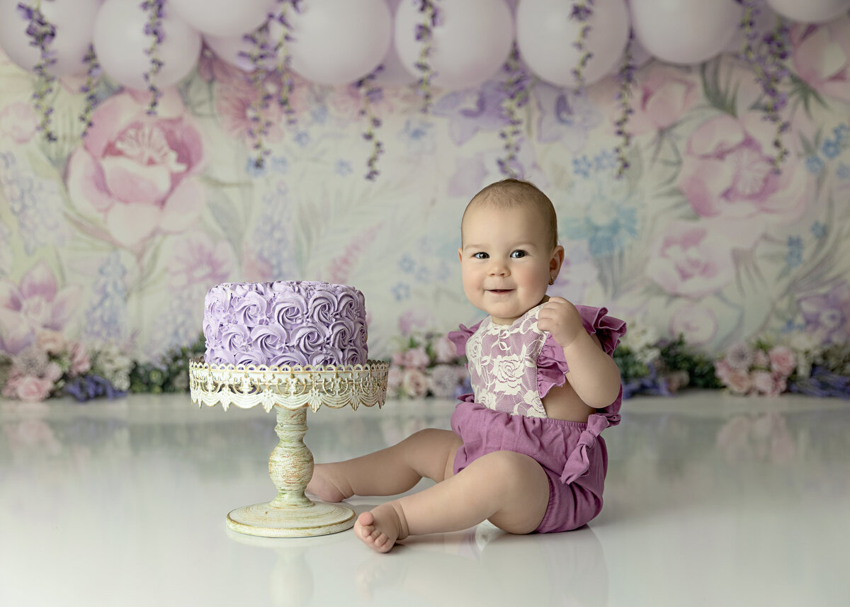 A toddler girl in a purple onesie sits in a floral themed studio for a birthday photo session