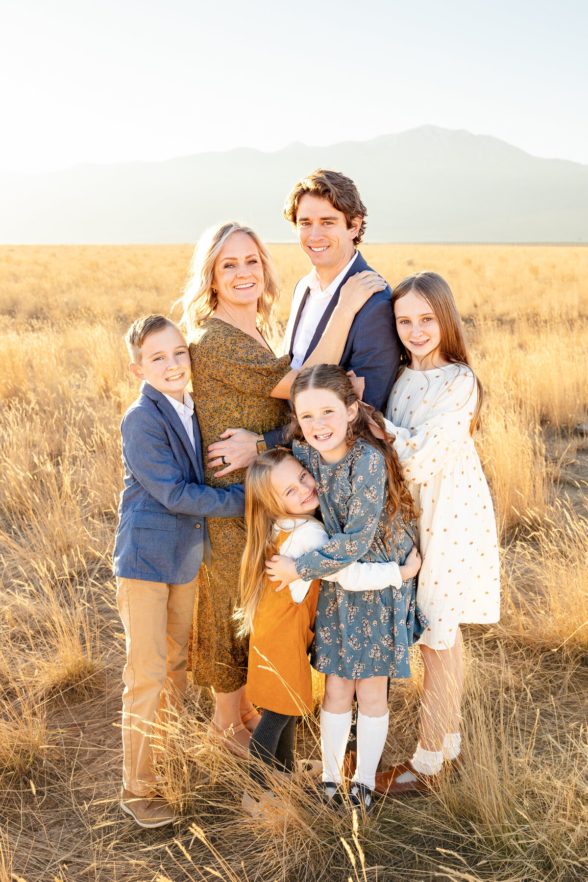 A loving family of six embraces each other in a golden field with mountains in the background, captured by Cassie Lee Photography.