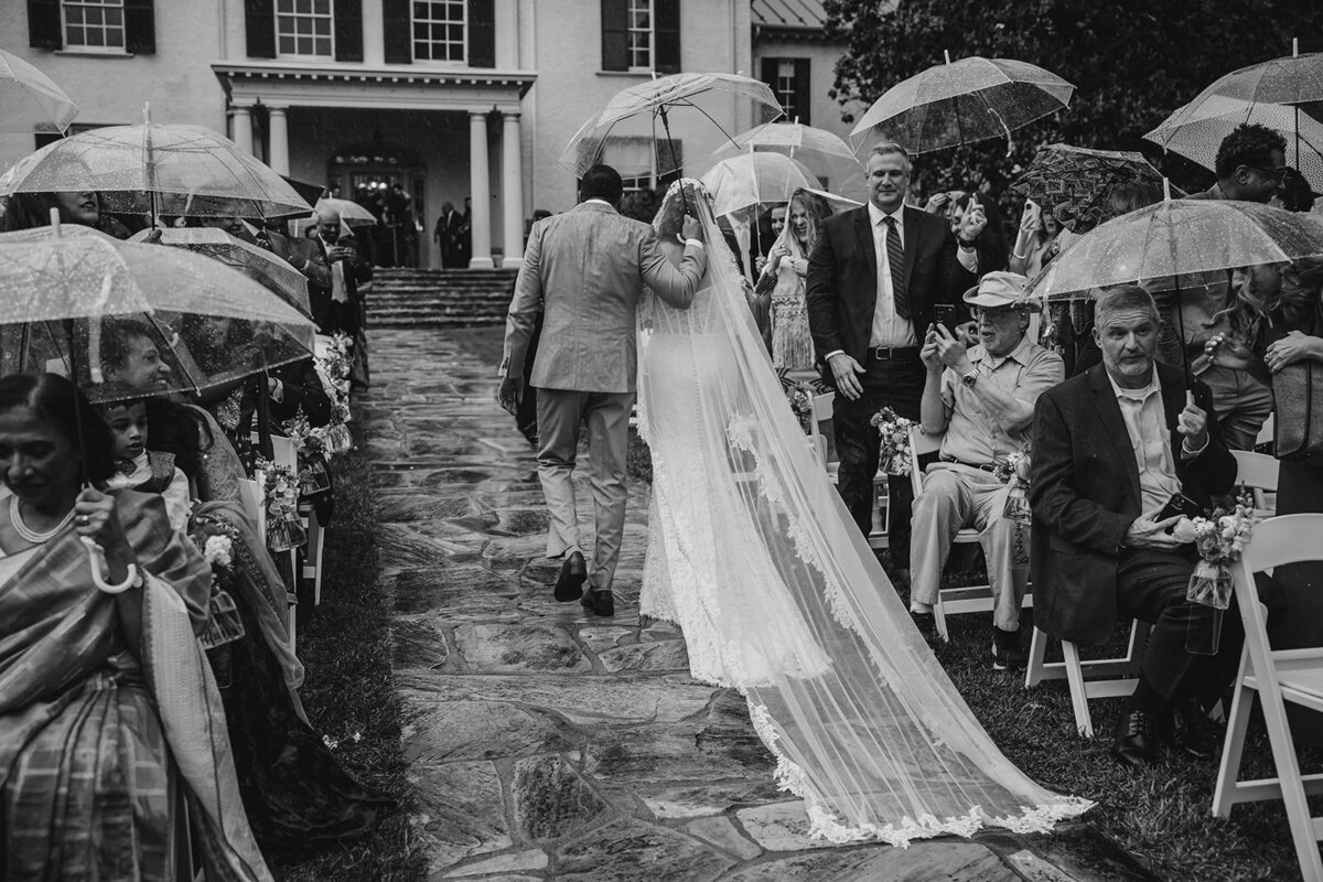 A couple walking back up the aisle in the rain under a clear umbrella 