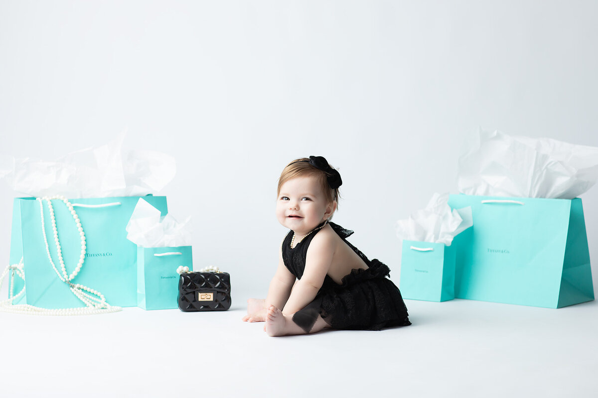 Dallas milestone photographer captures a stylish baby girl in a black lace outfit surrounded by Tiffany blue gift bags, pearls, and a mini designer purse for a glamorous one-year portrait.