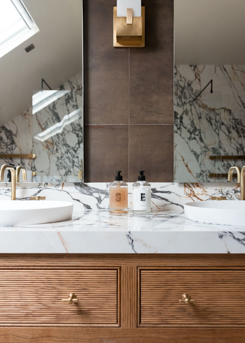 A modern bathroom, featuring a double sink vanity with marble countertops and wooden cabinets.The room is finished with dark tiled walls and light tiled floors.