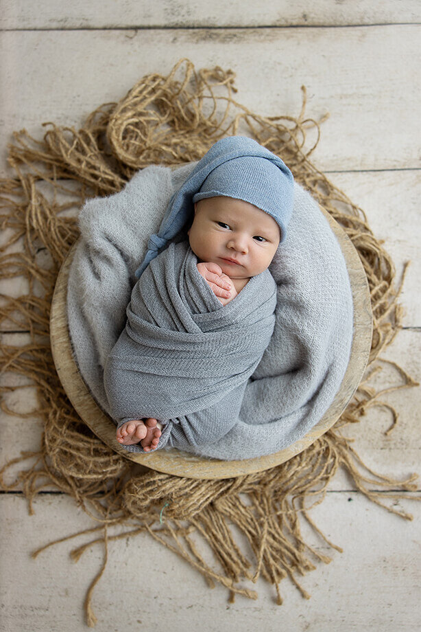 newborn boy is posed in a bowl with a soft blue criss cross wrap and he is wearing a soft blue sleepy hat.