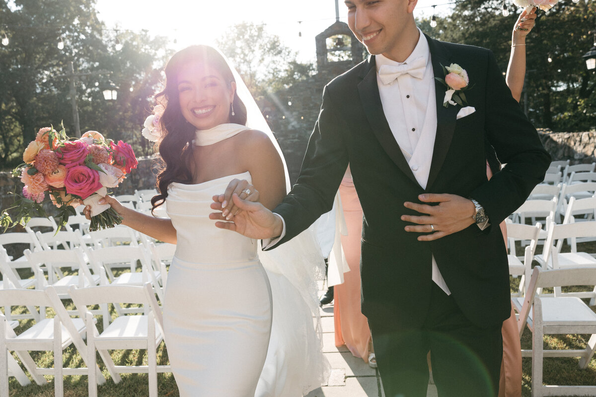 Joyful ceremony recessional as the bride carries a vibrant bouquet of garden roses, dahlias, and ranunculus while walking hand-in-hand with her groom under glowing afternoon light.
