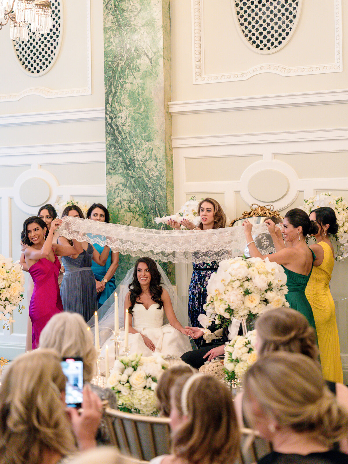 A bride sits on a chair in a grand room, surrounded by women holding a lace cloth above her. The women wear colorful dresses, and white floral arrangements adorn the scene. The atmosphere is elegant and festive.