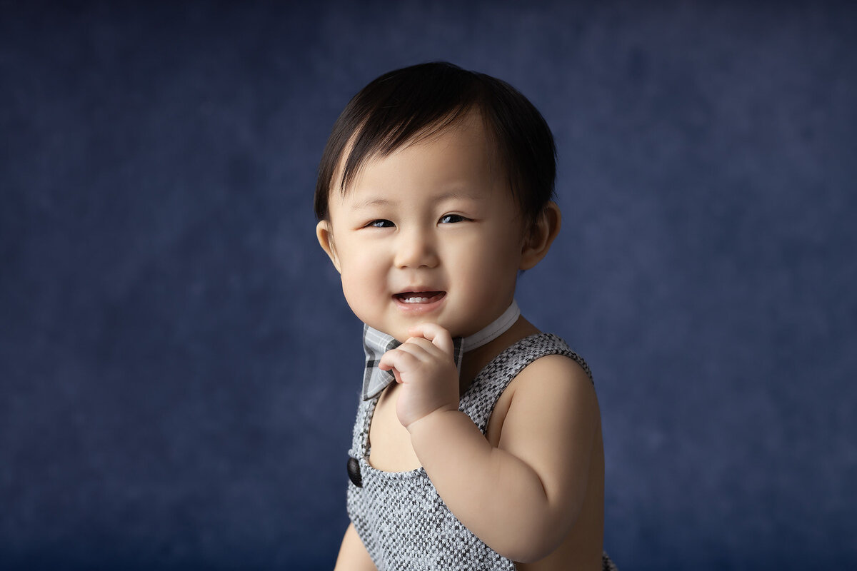 A smiling baby in a bowtie and overalls playfully touches their bowtie in front of a blue background.