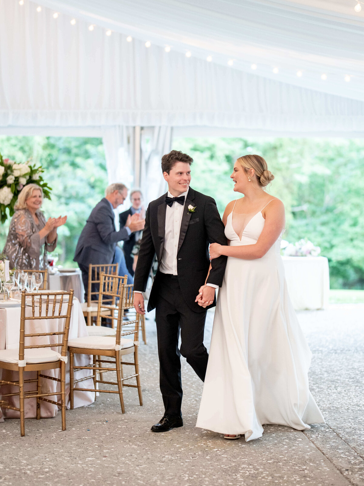 A bride and groom walk hand in hand under a white tent. The bride wears a flowing white dress, and the groom is in a black tuxedo. Guests are applauding around them, seated at tables with gold chairs and floral decorations.