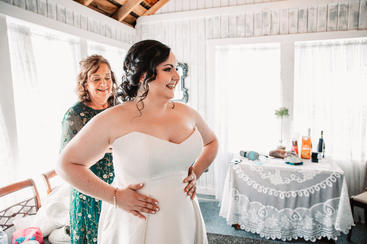 A smiling bride gets buttoned into her dress by mom in a white rustic room