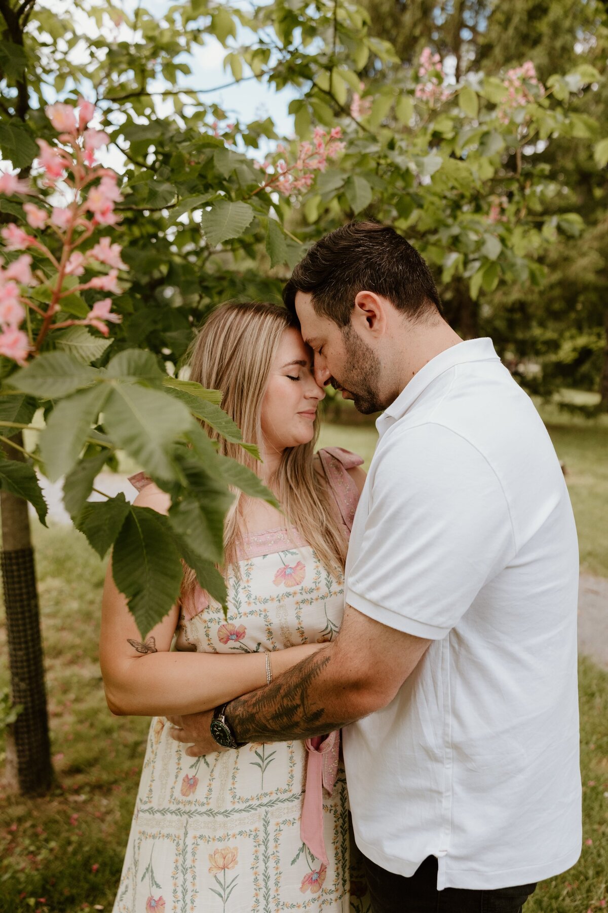 Couple shares an intimate forehead-to-forehead moment beneath pink flowering branches during a romantic spring engagement session in the park.