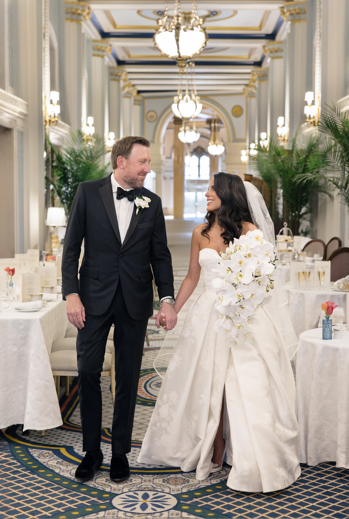 A bride and groom walk hand in hand down a lavishly decorated hall with elegant chandeliers and white floral arrangements. The bride wears a strapless white gown and holds a bouquet, while the groom is in a black tuxedo.