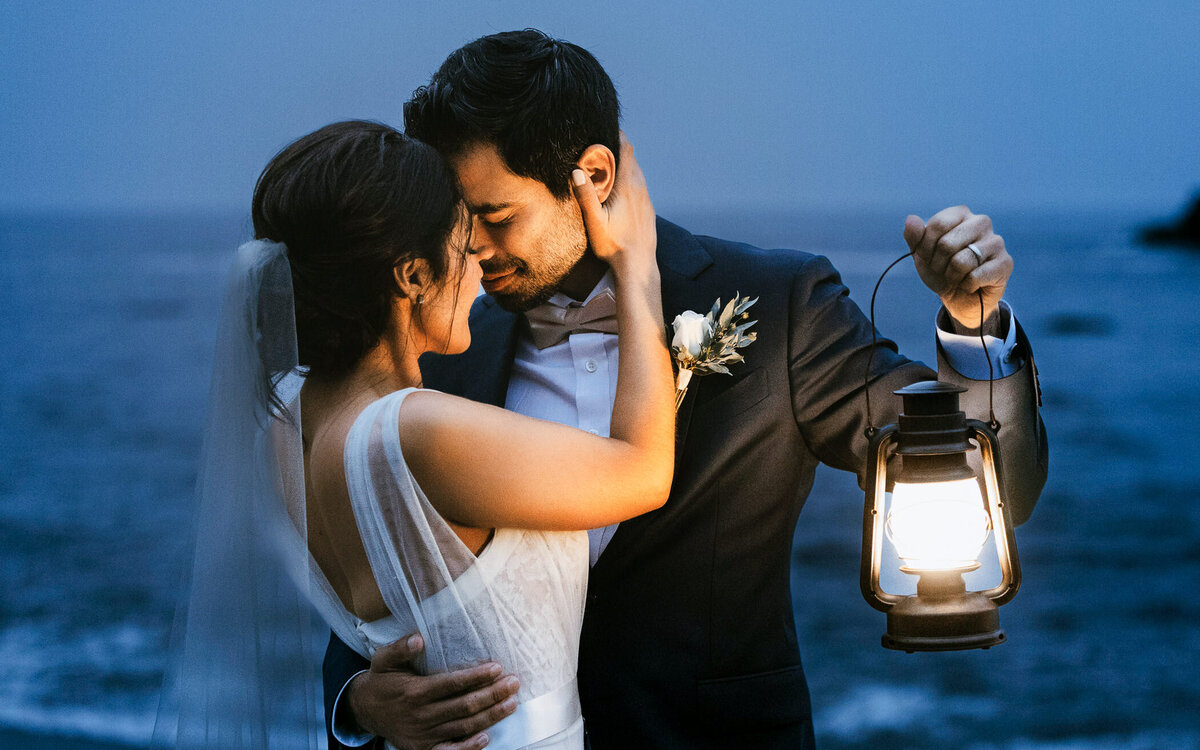 Bride and groom embrace by the ocean at dusk, holding a lantern for a cinematic wedding portrait.